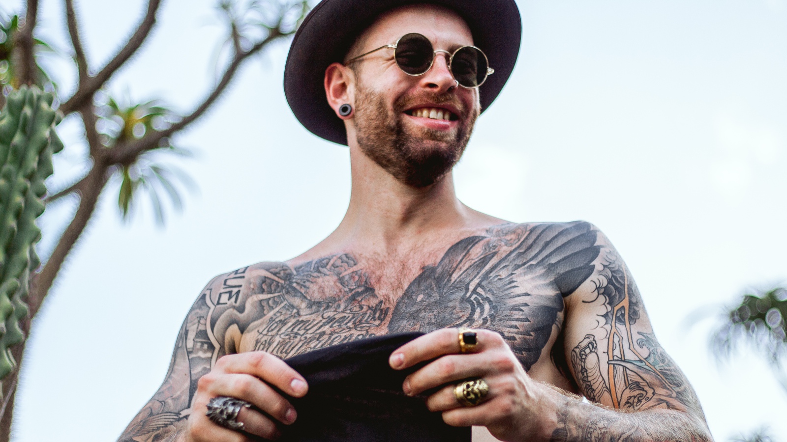 Мужчина с татуировками на теле в черной шляпе и очках