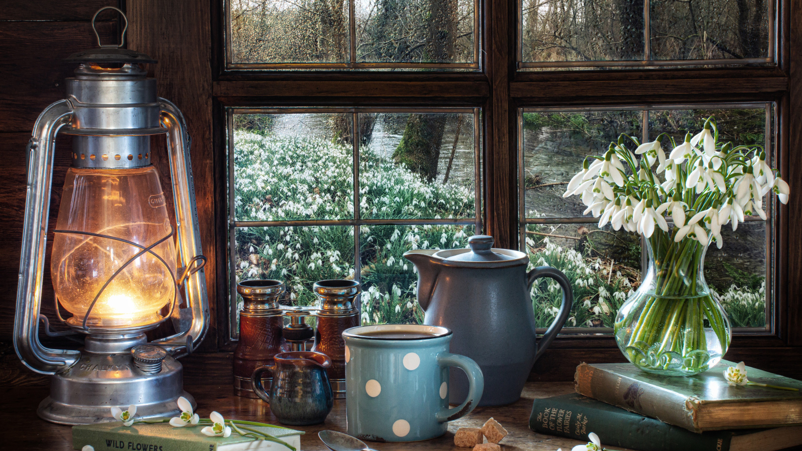 Керосиновая лампа на столе с кружкой, книгами и букетом подснежников у окна