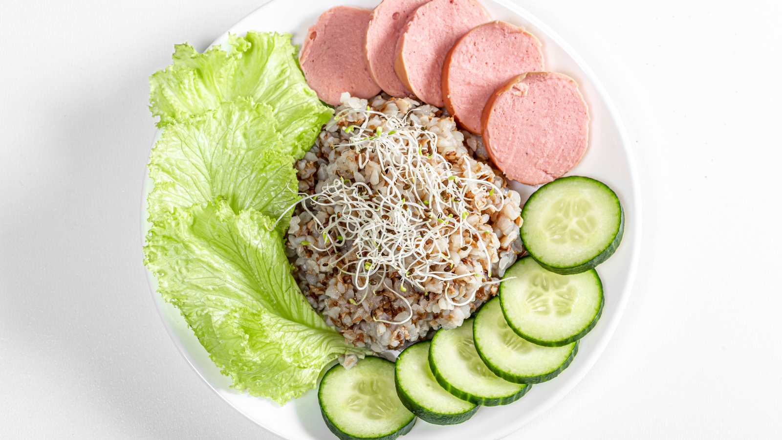 Гречка на тарелке с огурцом, колбасой и листьями салата на белом фоне