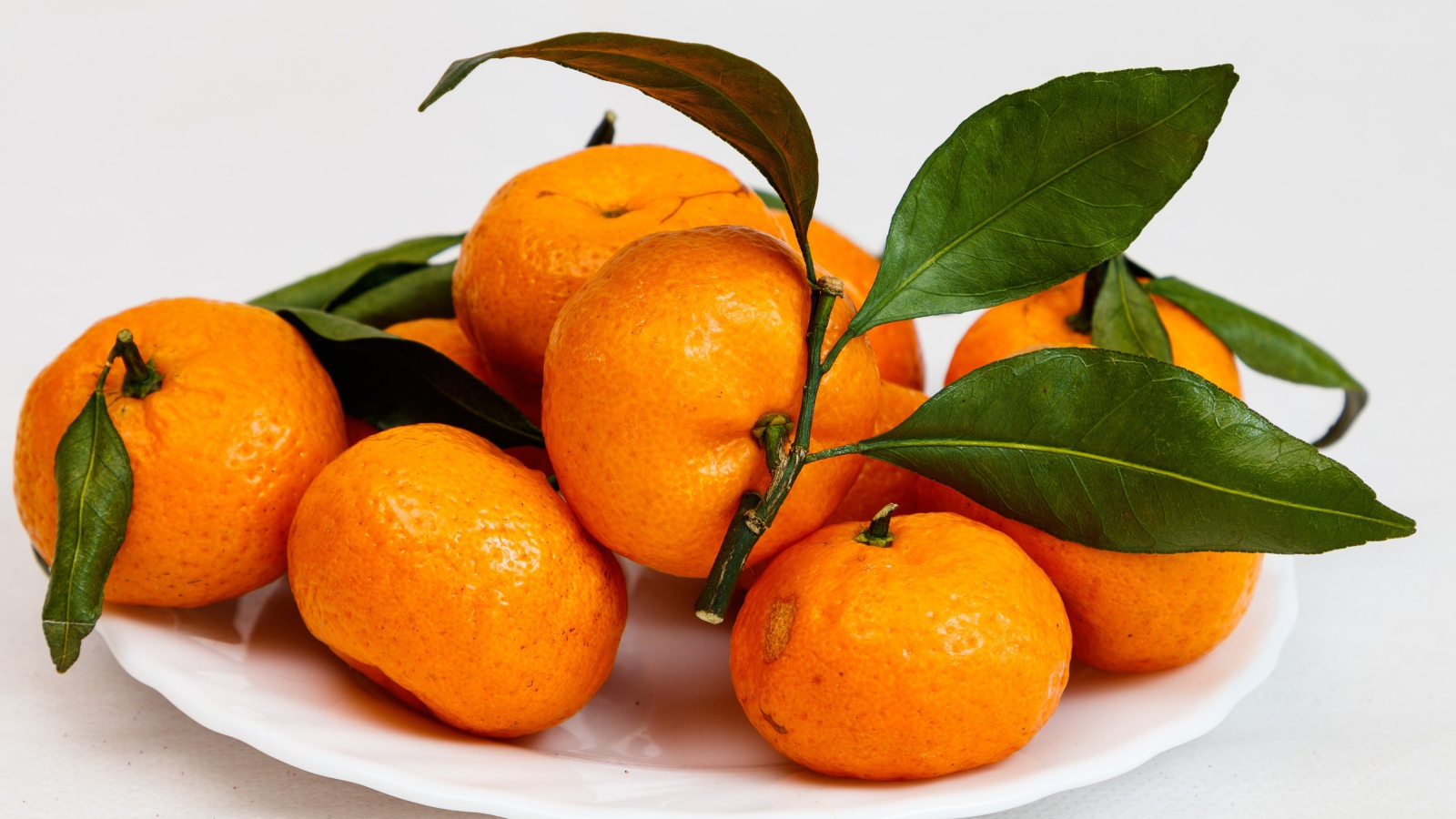 Свежие оранжевые мандарины с зелеными листьями на белой тарелке 