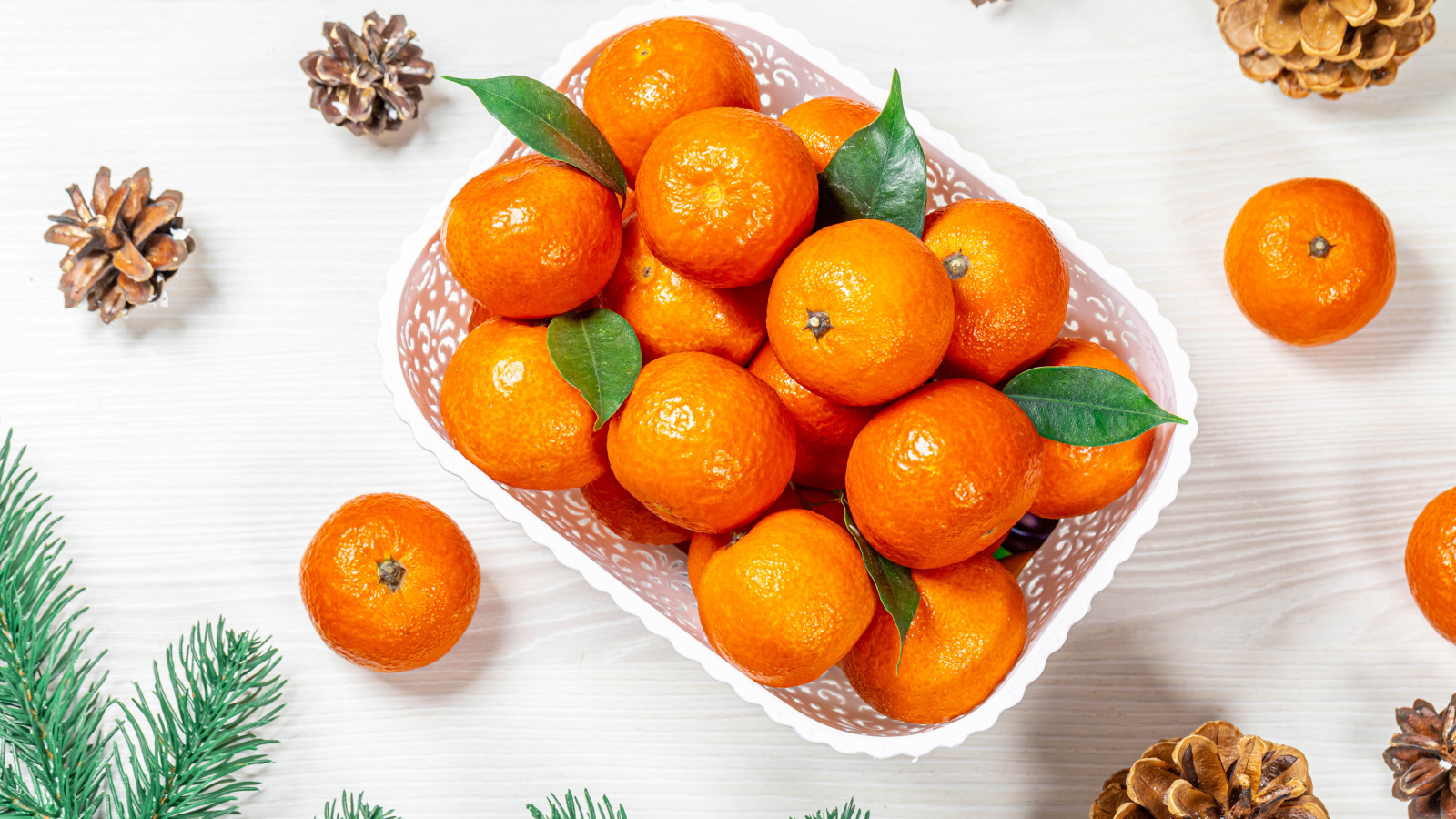 Много спелых мандаринов в белой корзинке на столе с шишками