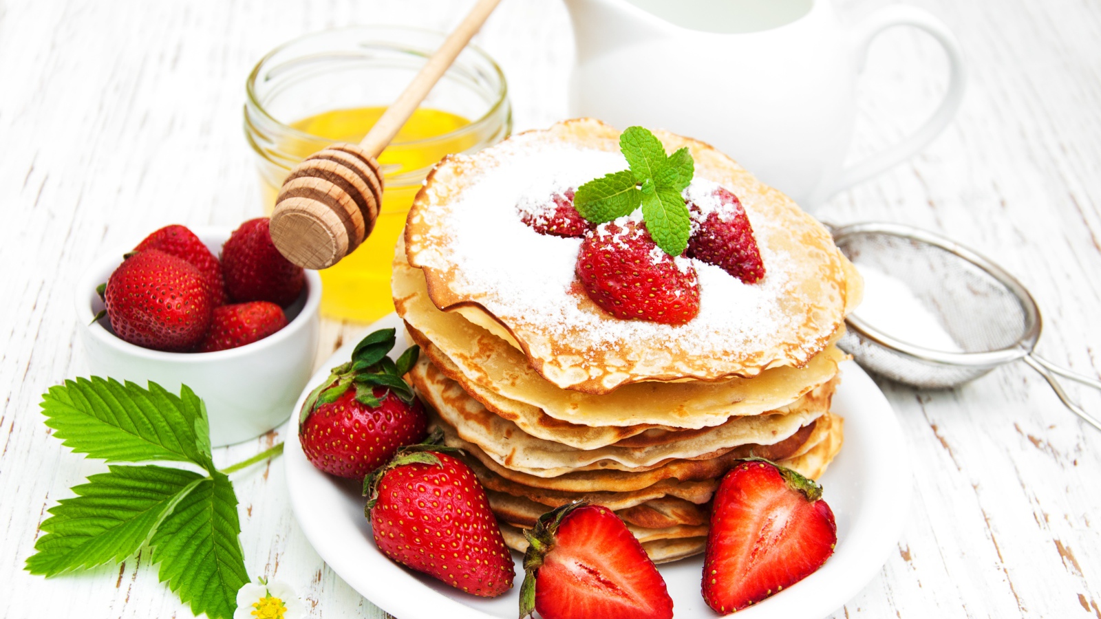 Тонкие блины с сахарной пудрой и ягодами клубники на столе с молоком и медом