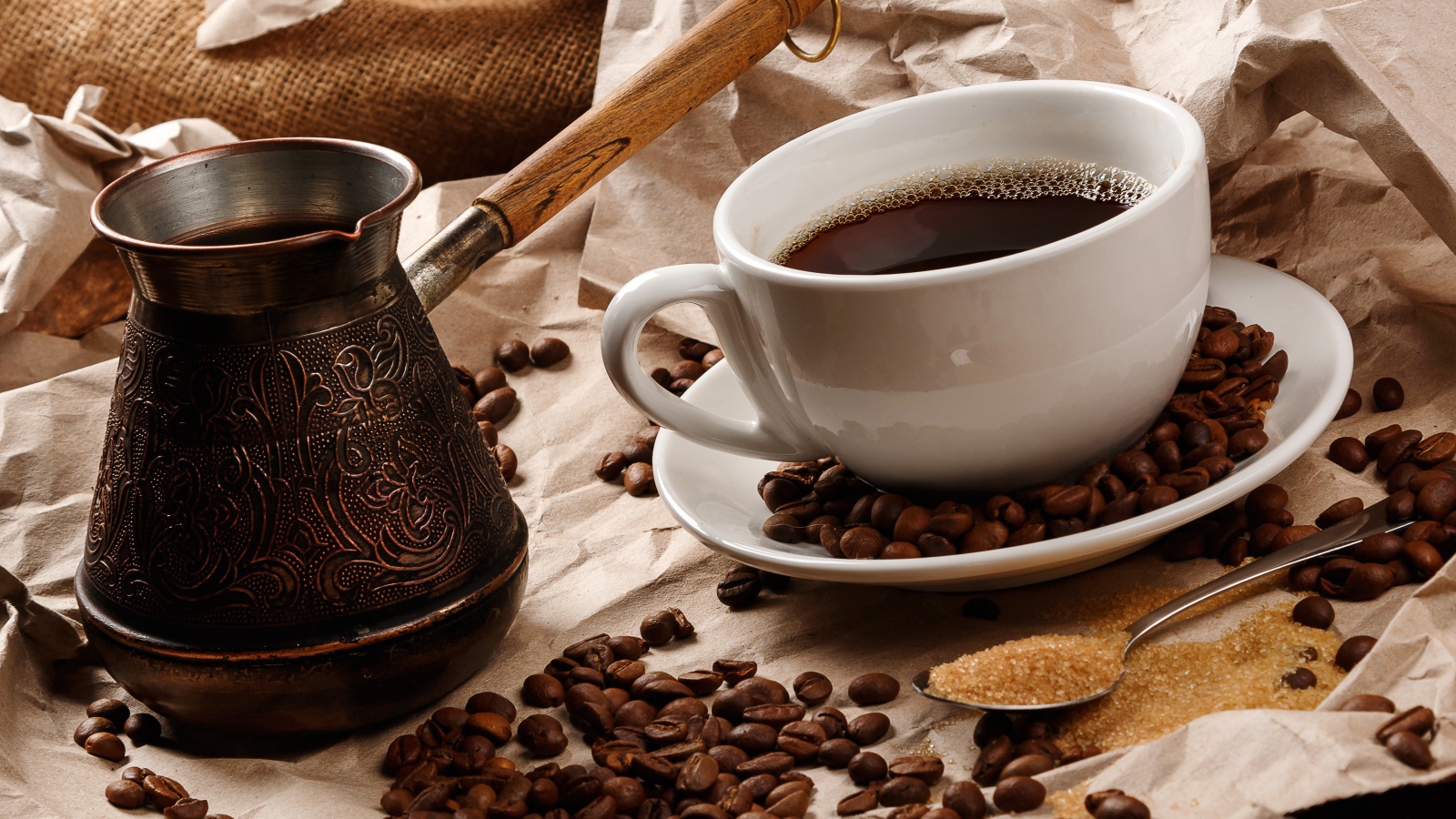 Чашка горячего кофе на столе с туркой, зернами и коричневым сахаром