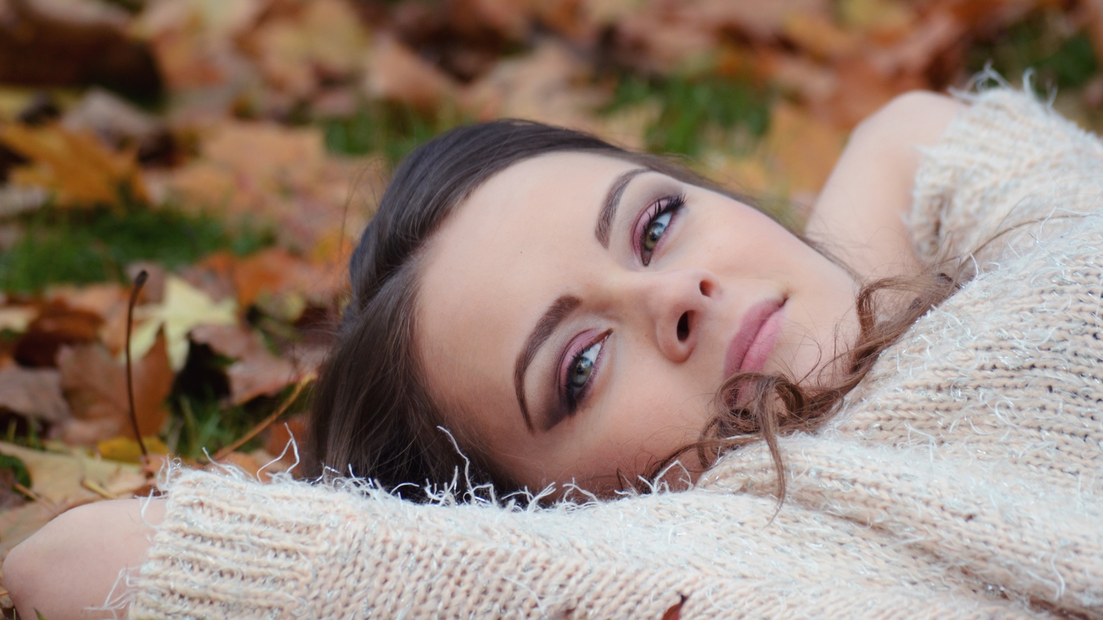 Красивая девушка в теплом свитере лежит на сухой листве 