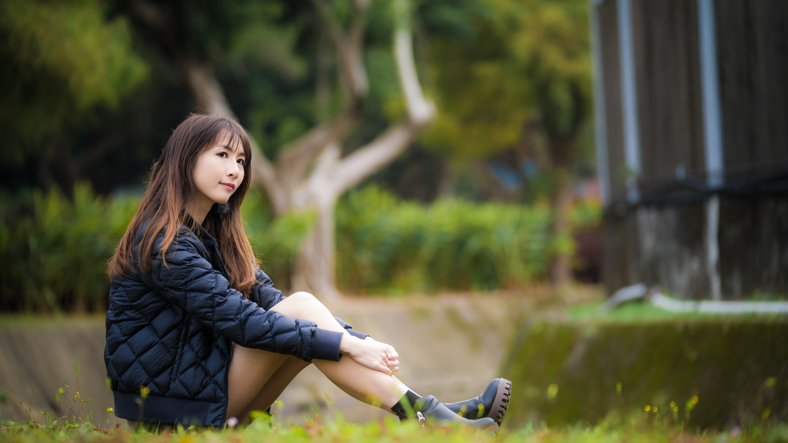Девушка азиатка в черной куртке сидит на траве