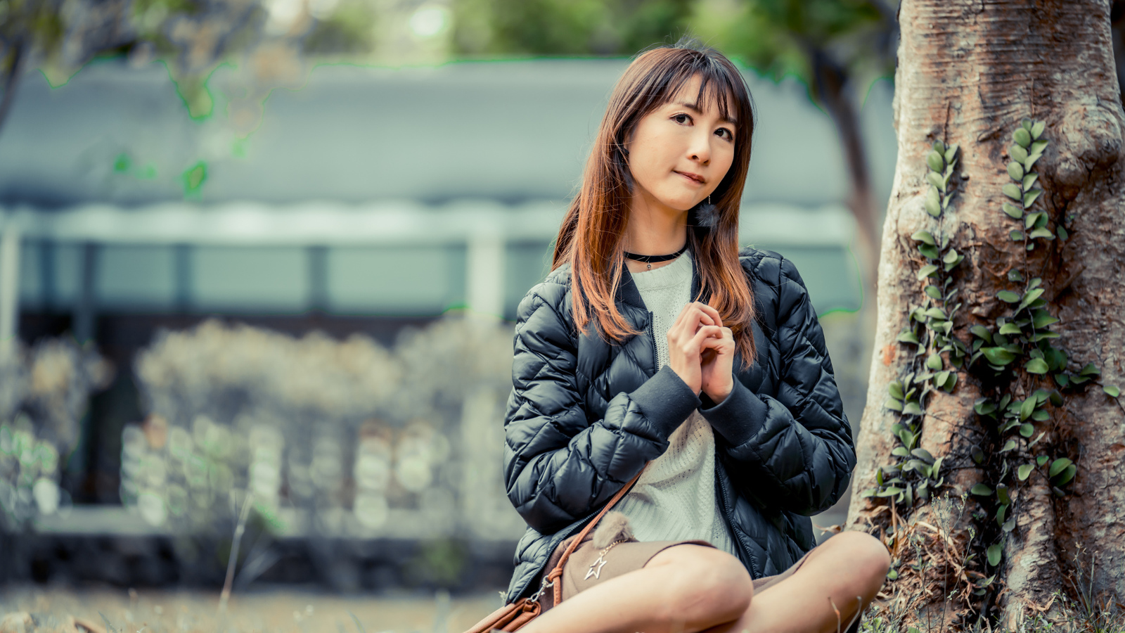 Девушка азиатка в черной куртке сидит под деревом
