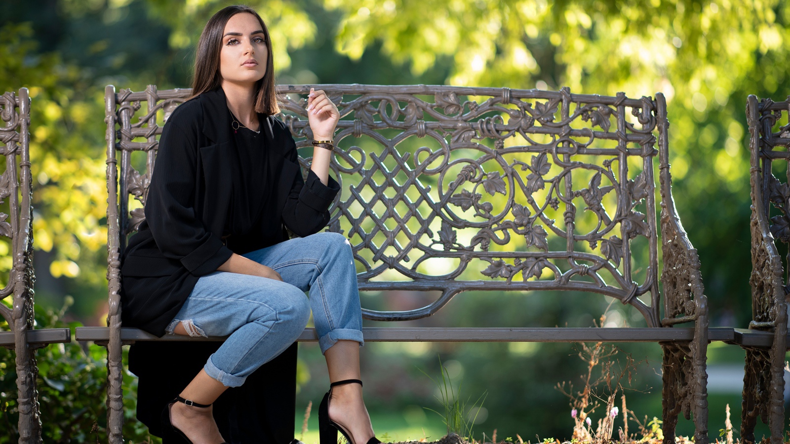 Красивая девушка в черном пальто сидит на лавочке в парке