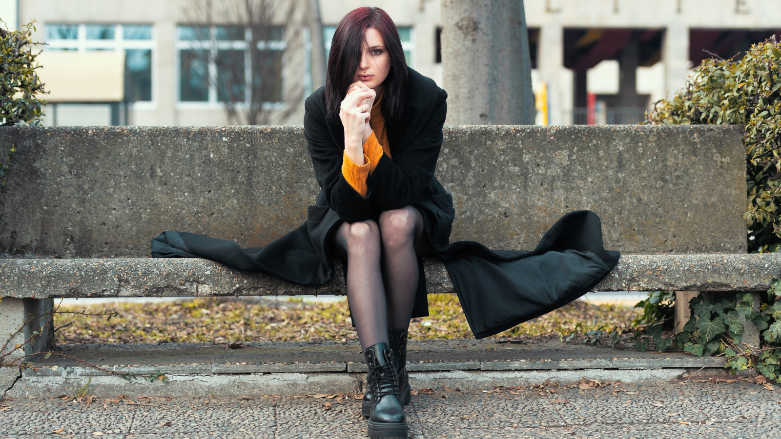 Девушка в черном пальто сидит на бетонной лавке 