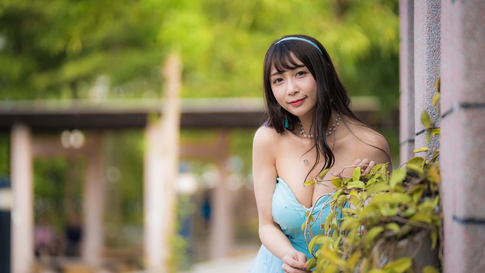 Улыбающаяся девушка азиатка в голубом платье у стены с растением 