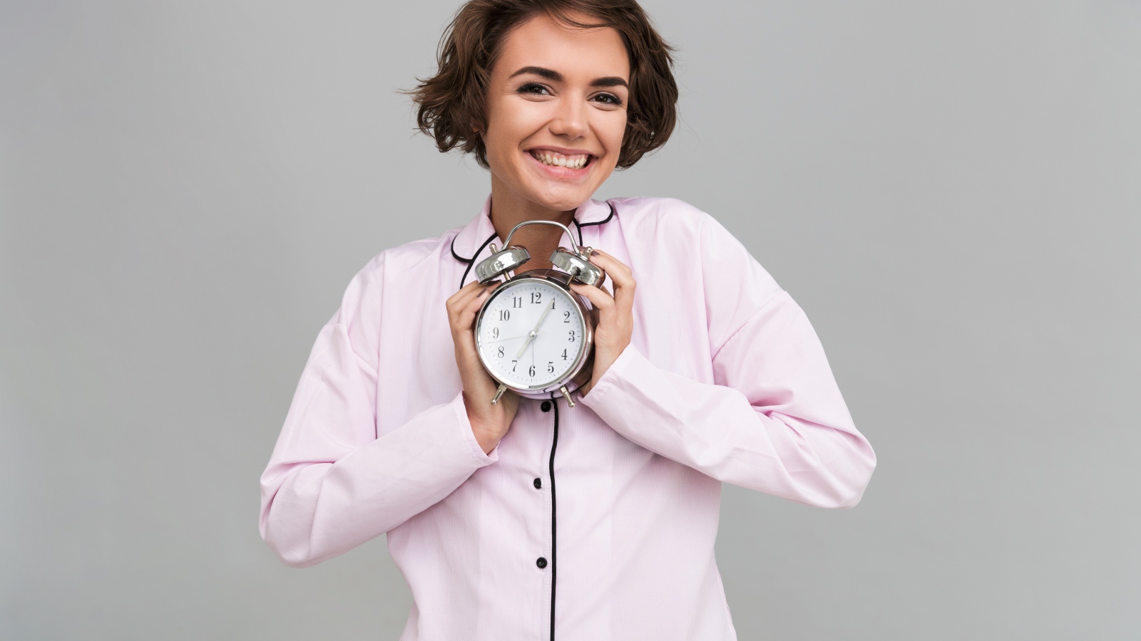 Улыбающаяся девушка в пижаме держит будильник 