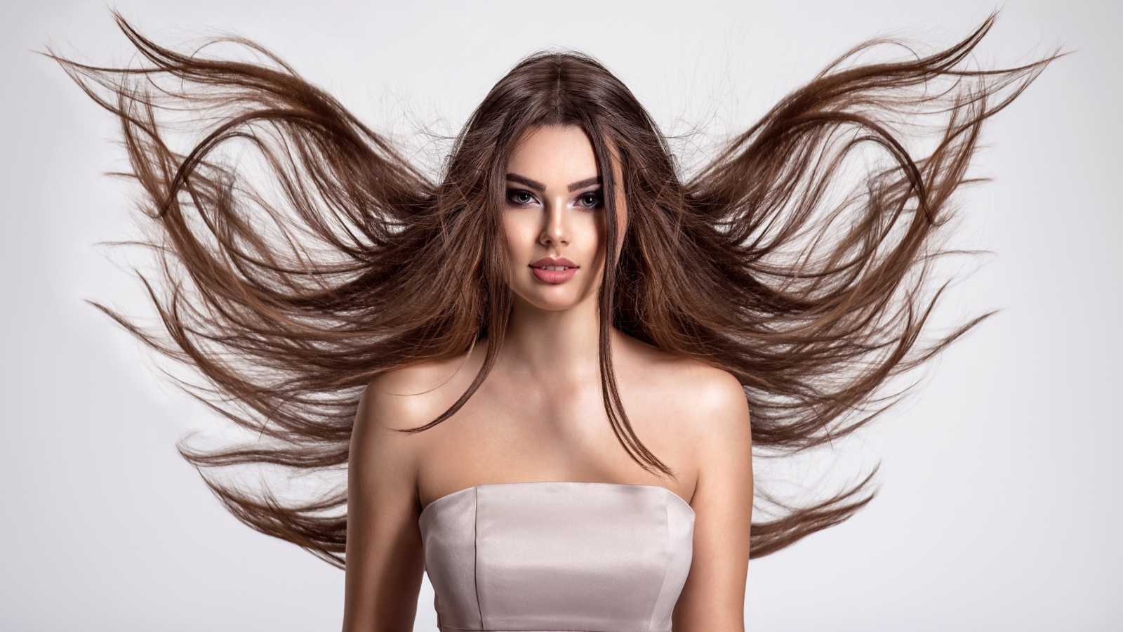 Ветер развивает волосы красивой девушке шатенке на сером фоне