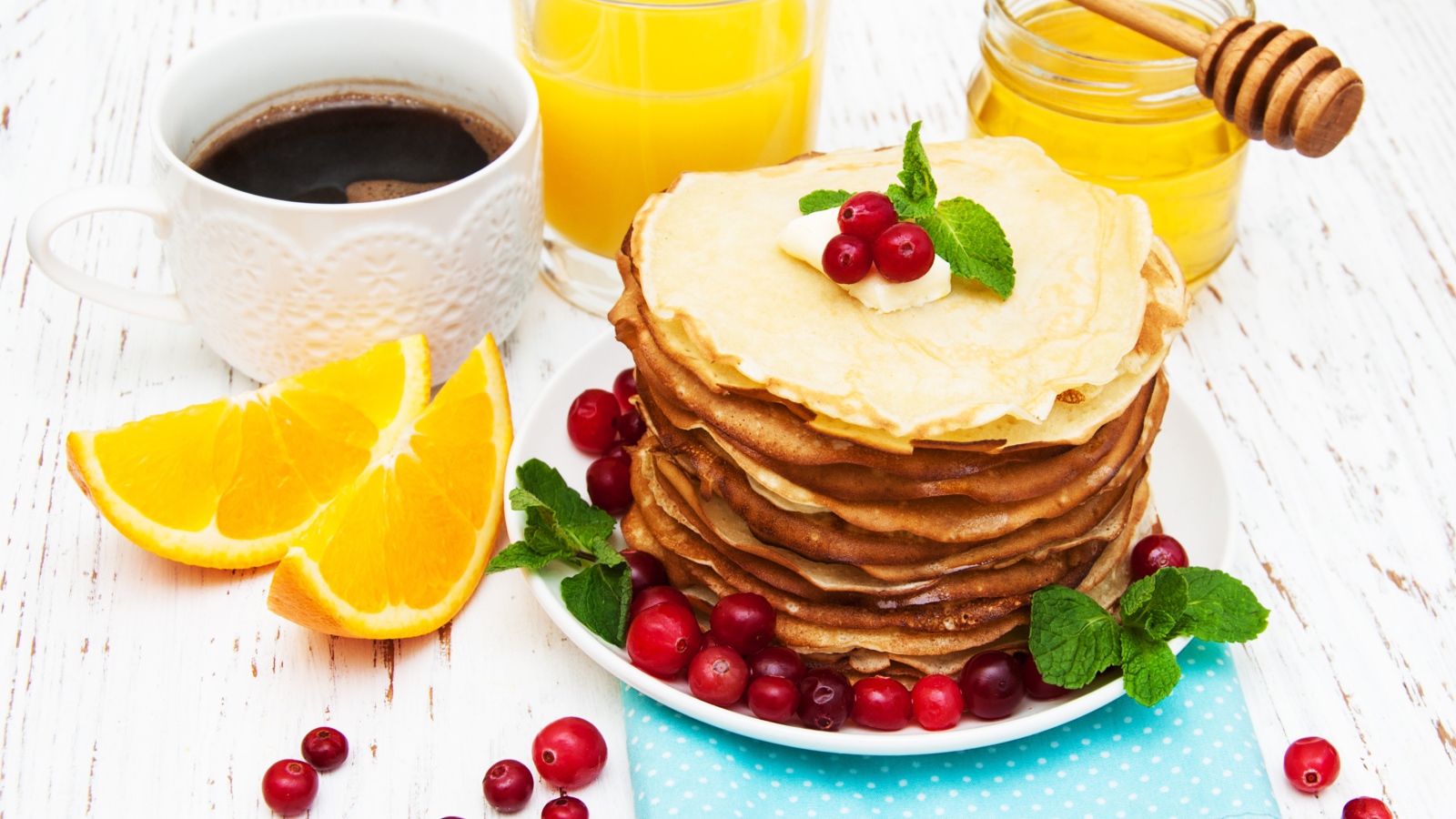 Тонкие румяные блинчики на столе с ягодами клюквы, апельсинами, медом и кофе