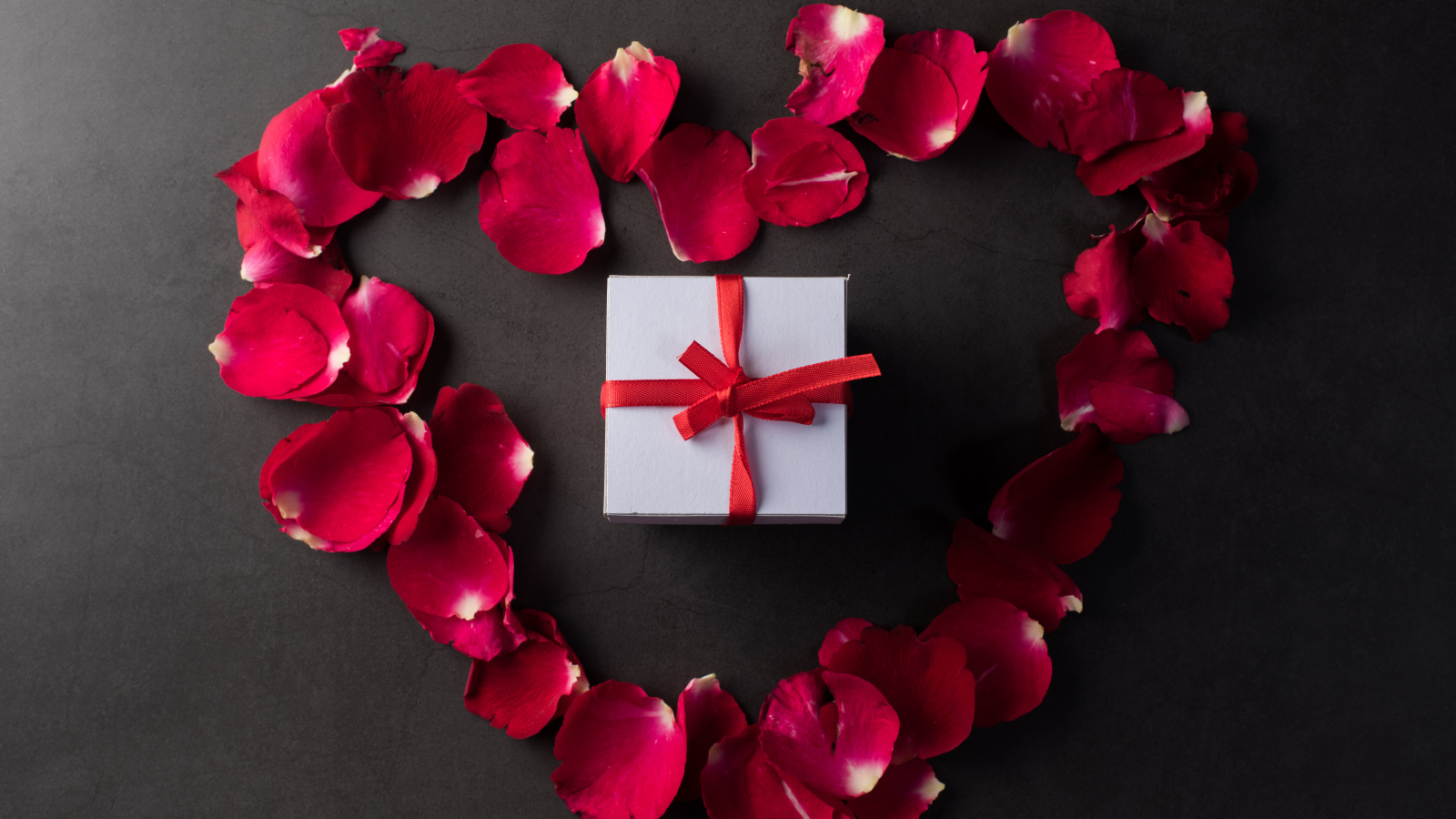 Сердце из красных лепестков розы с подарком на сером фоне