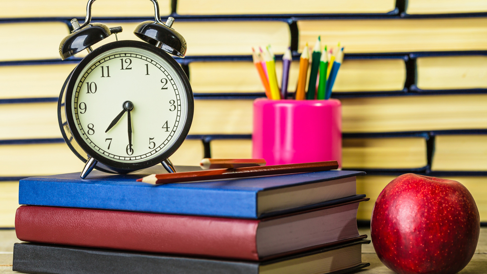 Будильник, книги, карандаши и яблоко на столе у стены 