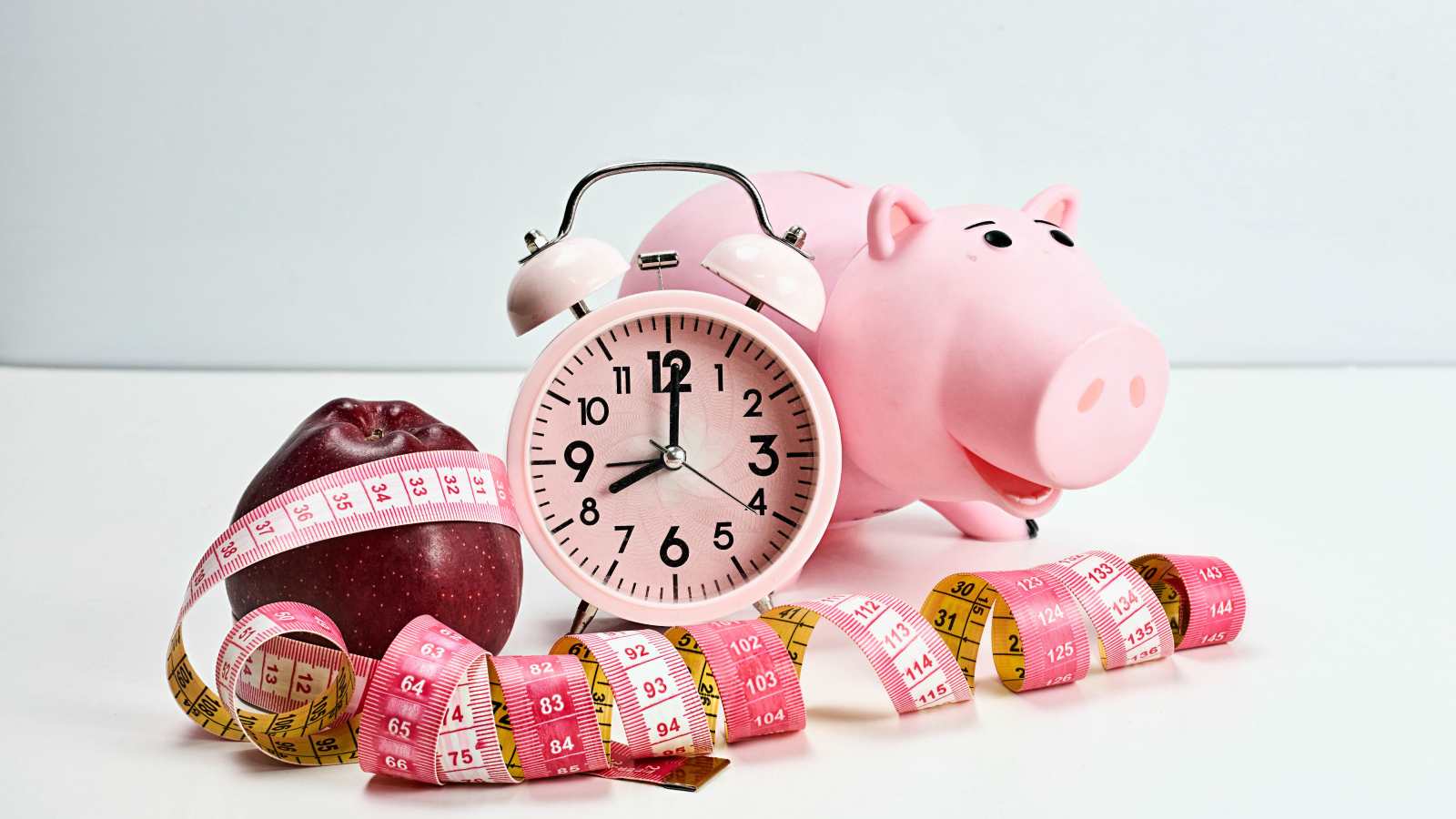 Будильник, свинья копилка, яблоко и сантиметр на белом фоне