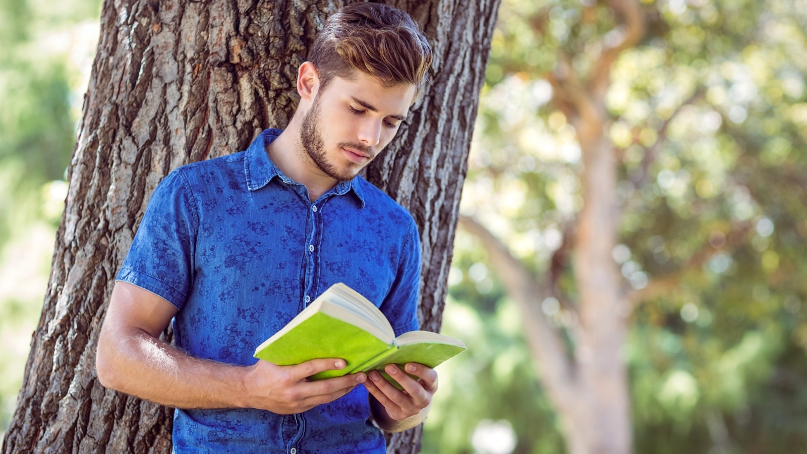 Красивый молодой мужчина стоит с книгой под деревом 