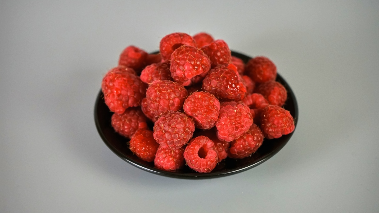 Спелая красная малина в черной тарелке на сером фоне
