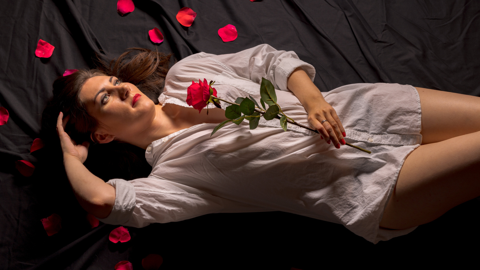 Девушка в белой рубашке с розой в руках лежит на постели