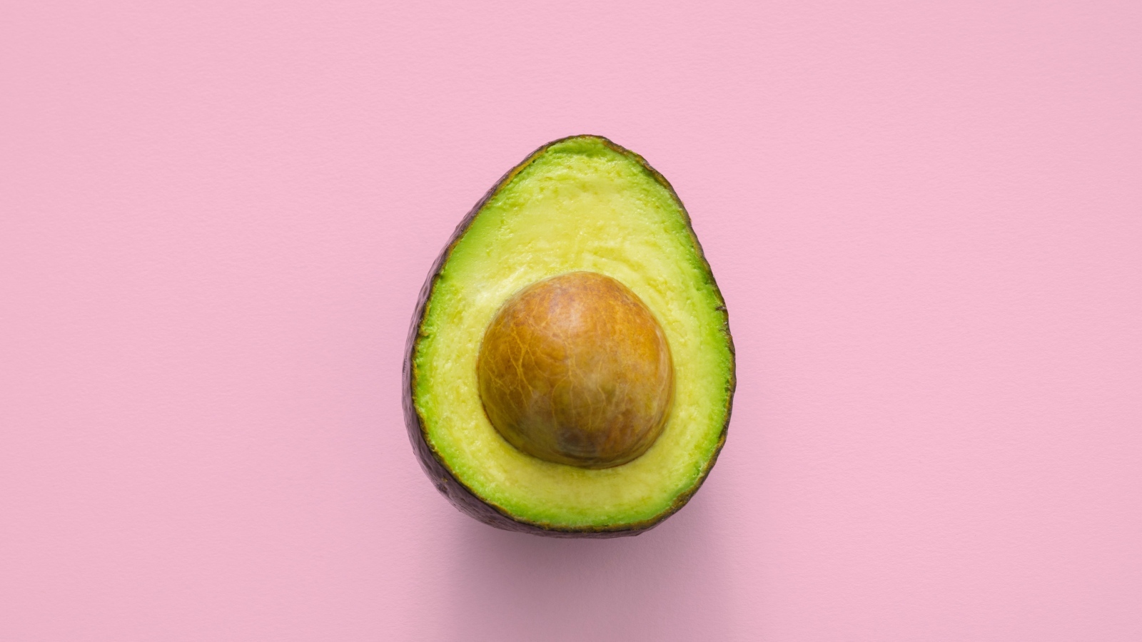 Половина авокадо с большой косточкой на розовом фоне