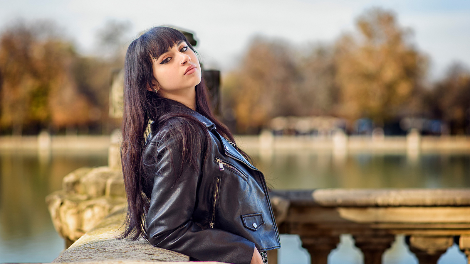 Девушка в черной куртке стоит на мосту у воды