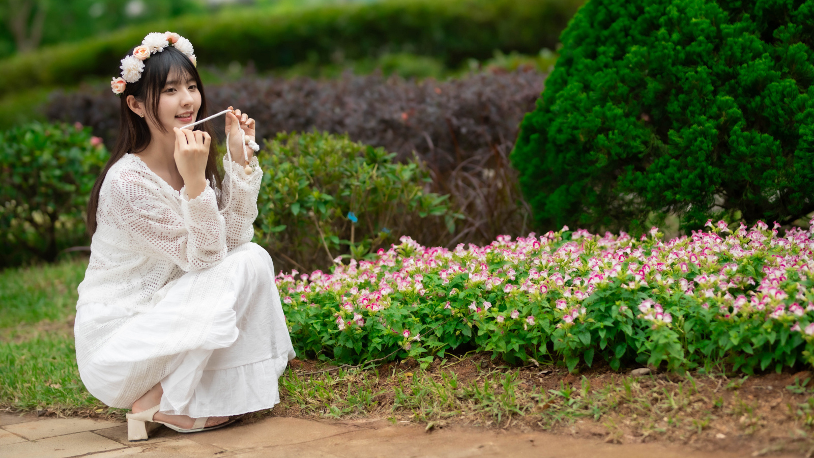 Милая азиатка в белом наряде сидит у клумбы с цветами