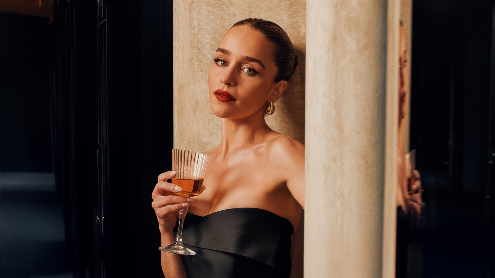 Актриса Эмилия Кларк с бокалом стоит у стены