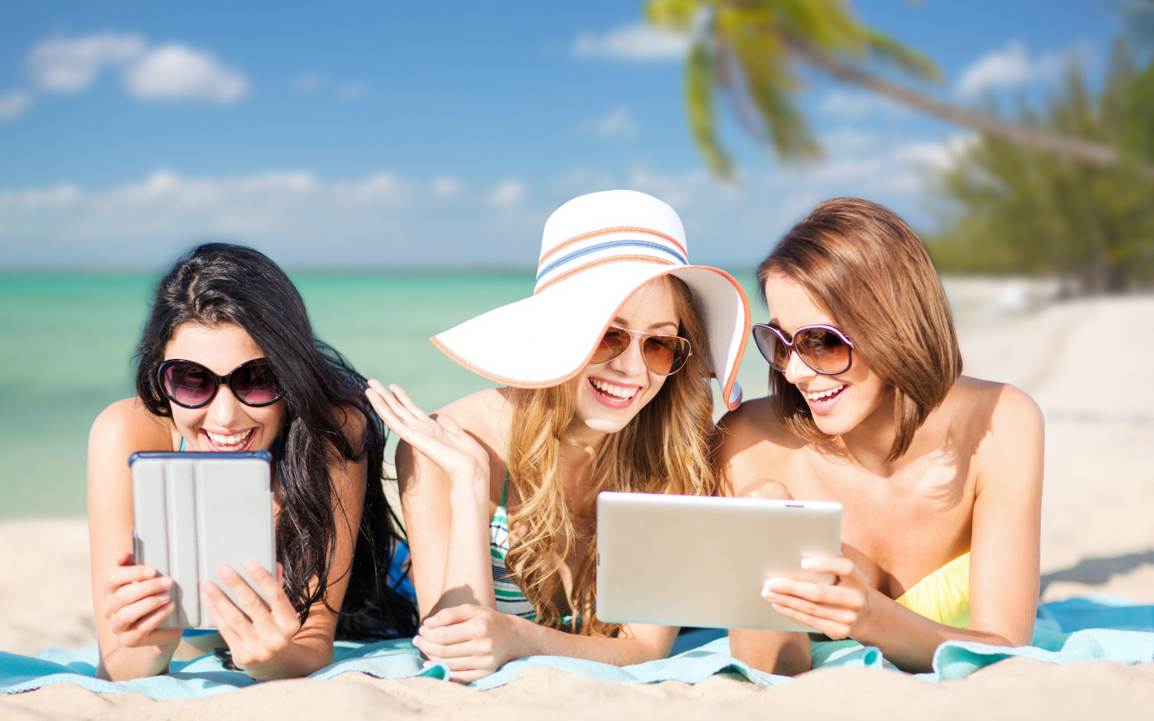 Три улыбающиеся девушки загорают на пляже с планшетами в руках