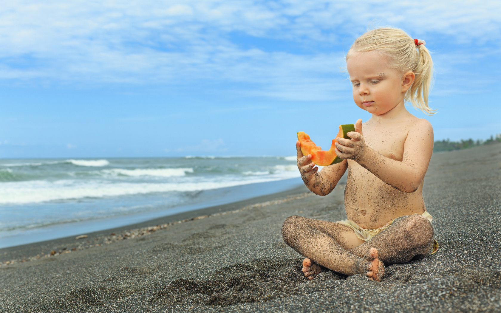 Маленькая девочка сидит на песке у моря с кусочком дыни