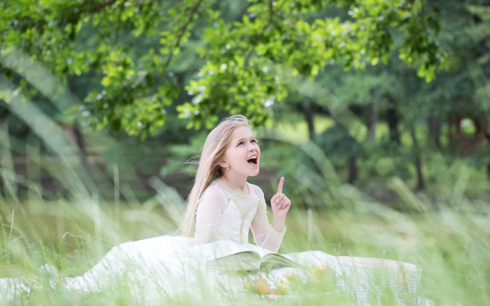 Красивая маленькая девочка в белом платье с книгой сидит на траве