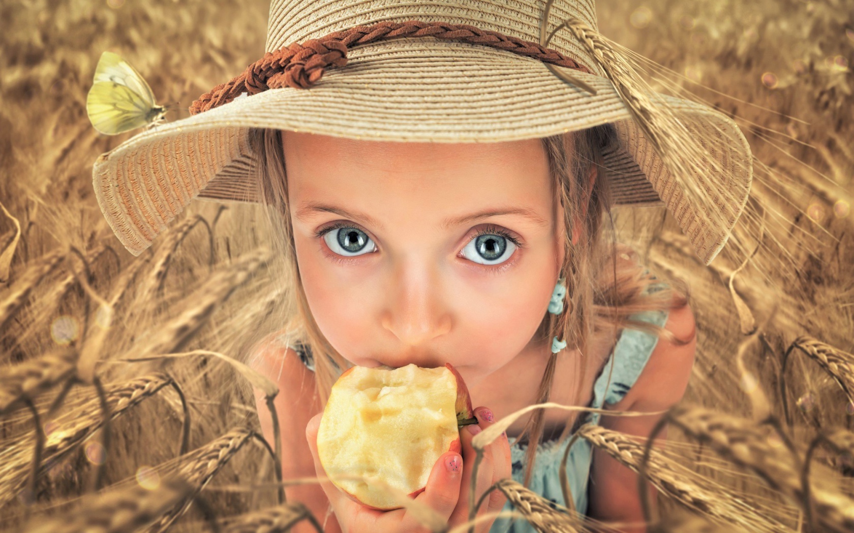 Маленькая голубоглазая девочка в шляпе ест яблоко