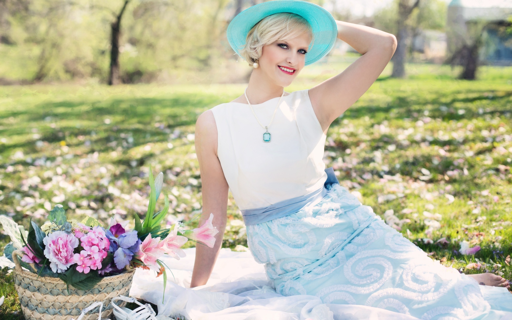 Красивая блондинка в голубой шляпке сидит в парке на траве