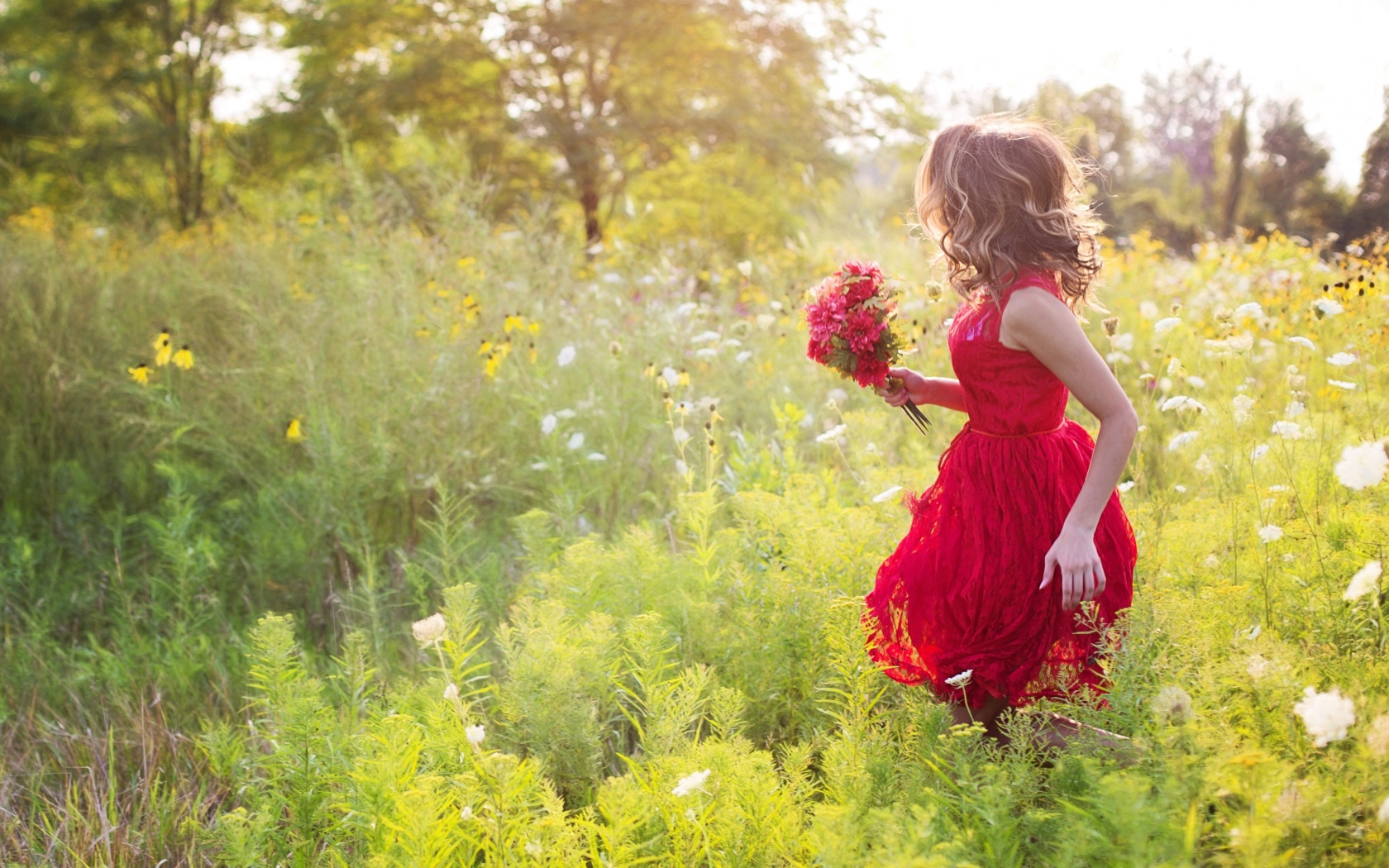 Красивая девушка в красном платье гуляет с букетом в руках по траве