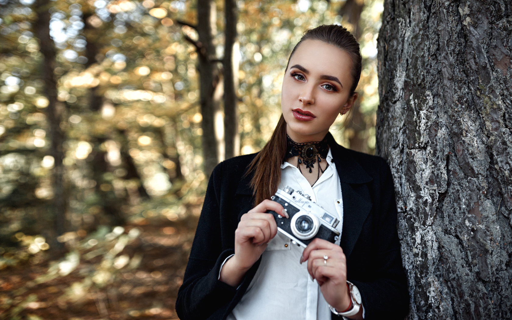 Красивая девушка с фотоаппаратом в руках стоит у дерева