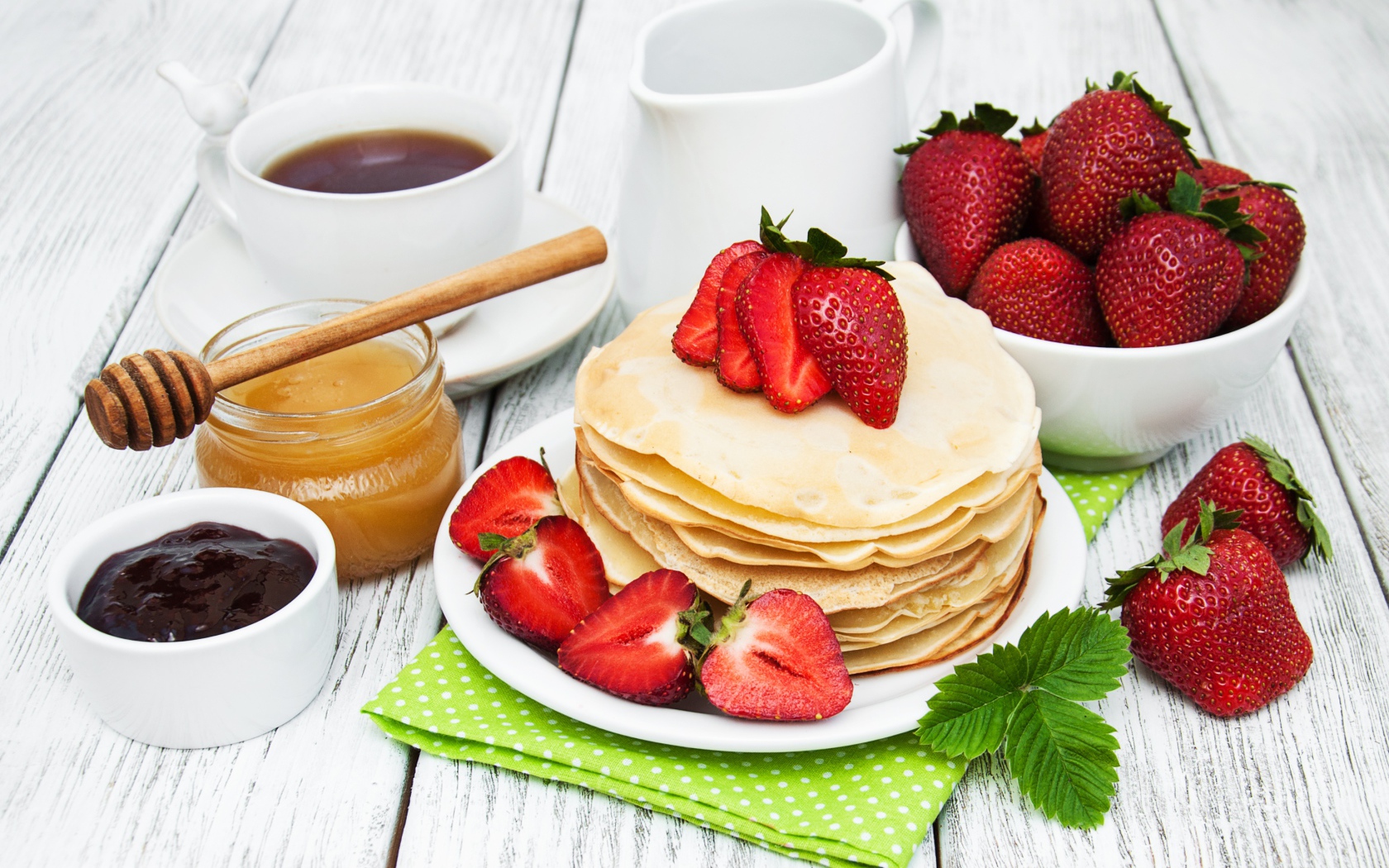 Тонкие блины на столе с ягодами клубники, медом, чаем и джемом