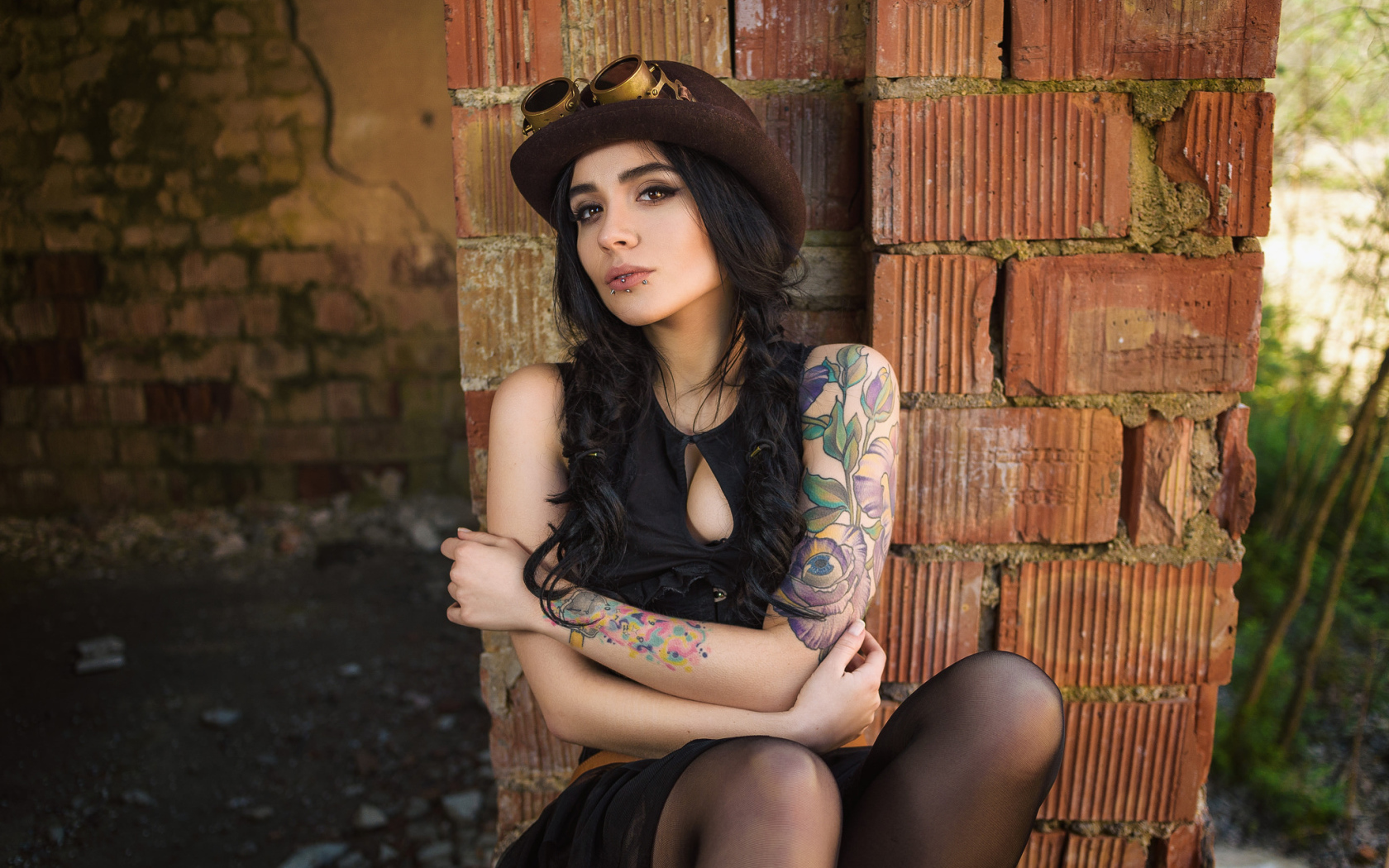 Девушка в шляпе с татуировками на руке сидит у стены