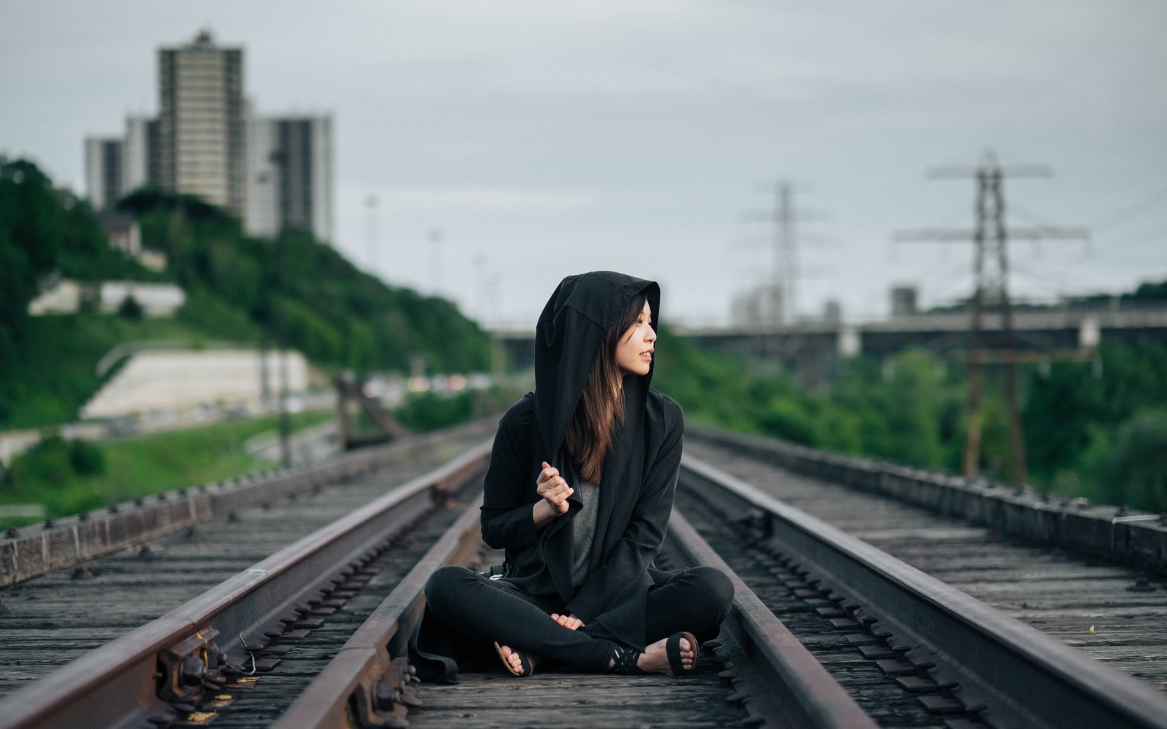 Девушка азиатка в черном балахоне сидит на рельсах 