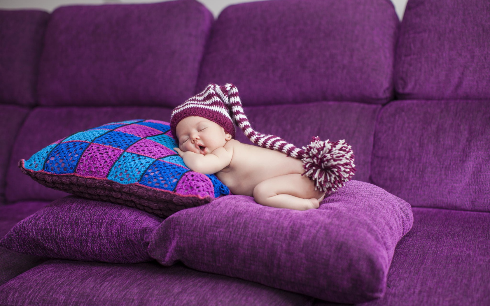 Маленький ребенок в большой шапке спит на фиолетовых подушках