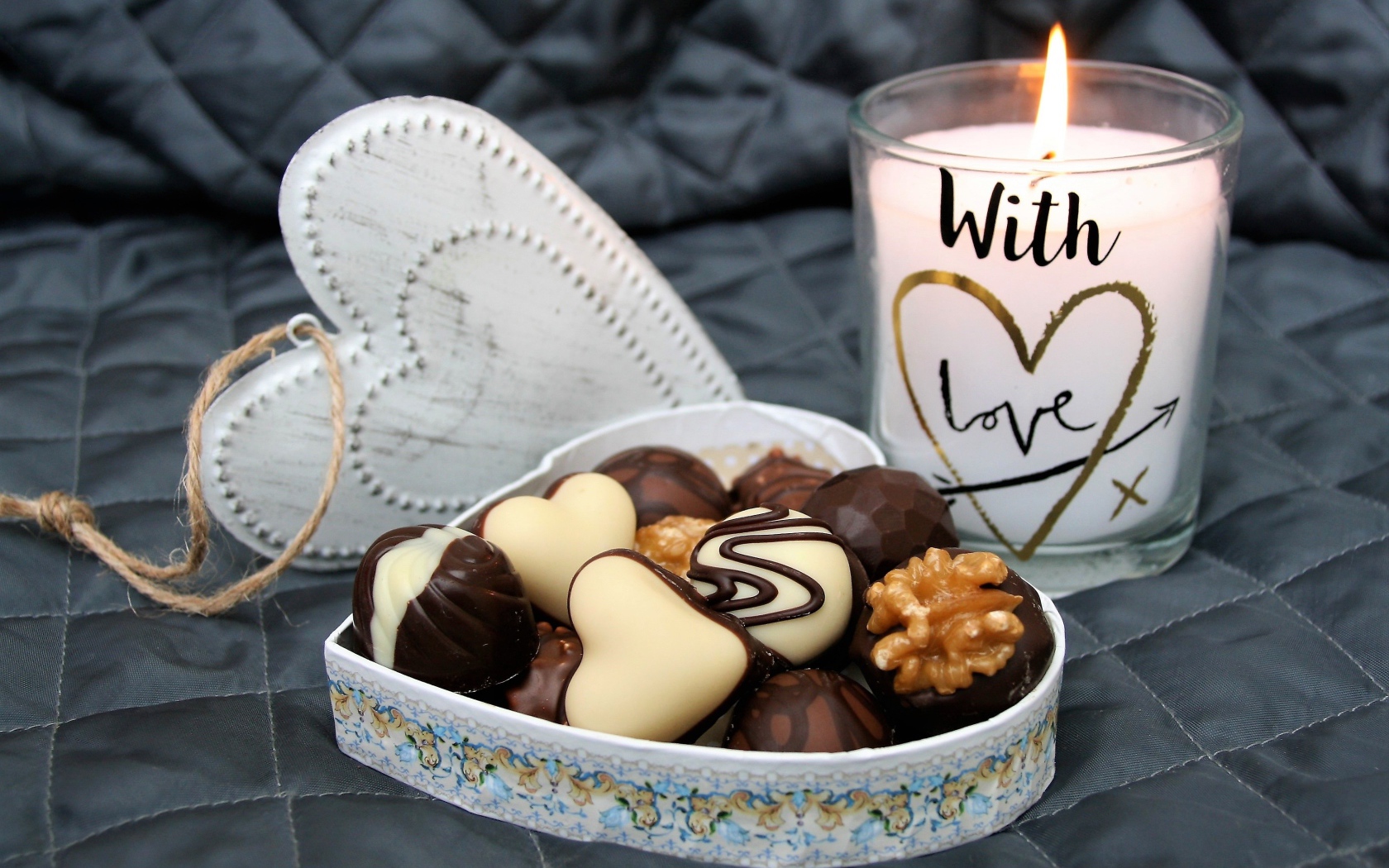 Chocolates in a heart shaped box on a bed with a lit candle