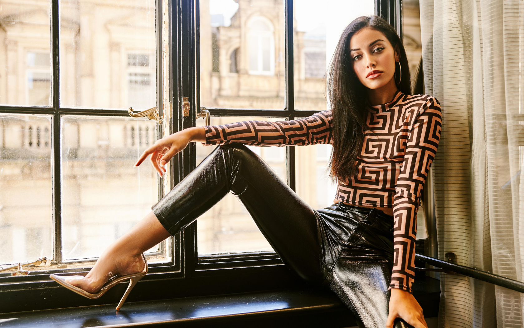 Красивая брюнетка в черных брюках сидит на окне 