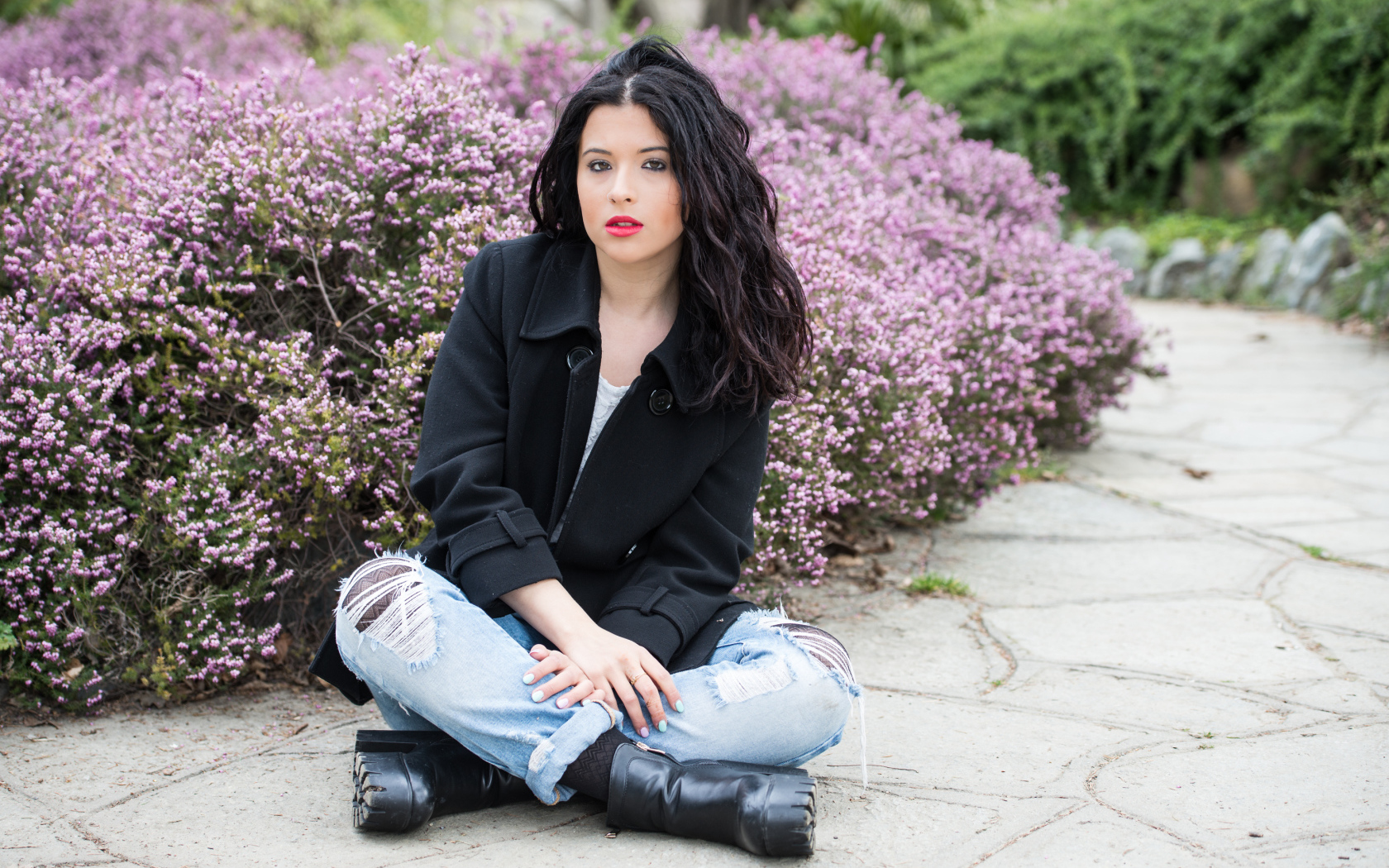 Красивая девушка в черном пальто сидит на земле у кустов