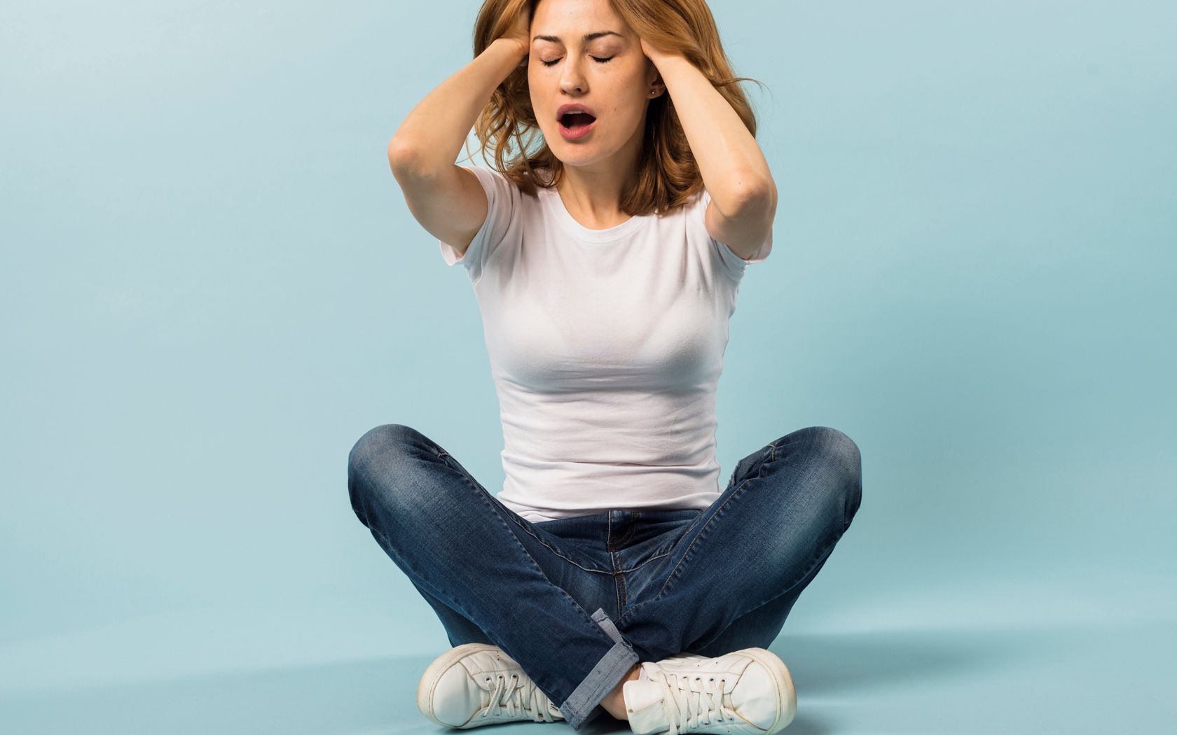 Девушка с руками в волосах сидит в джинсах на голубом фоне
