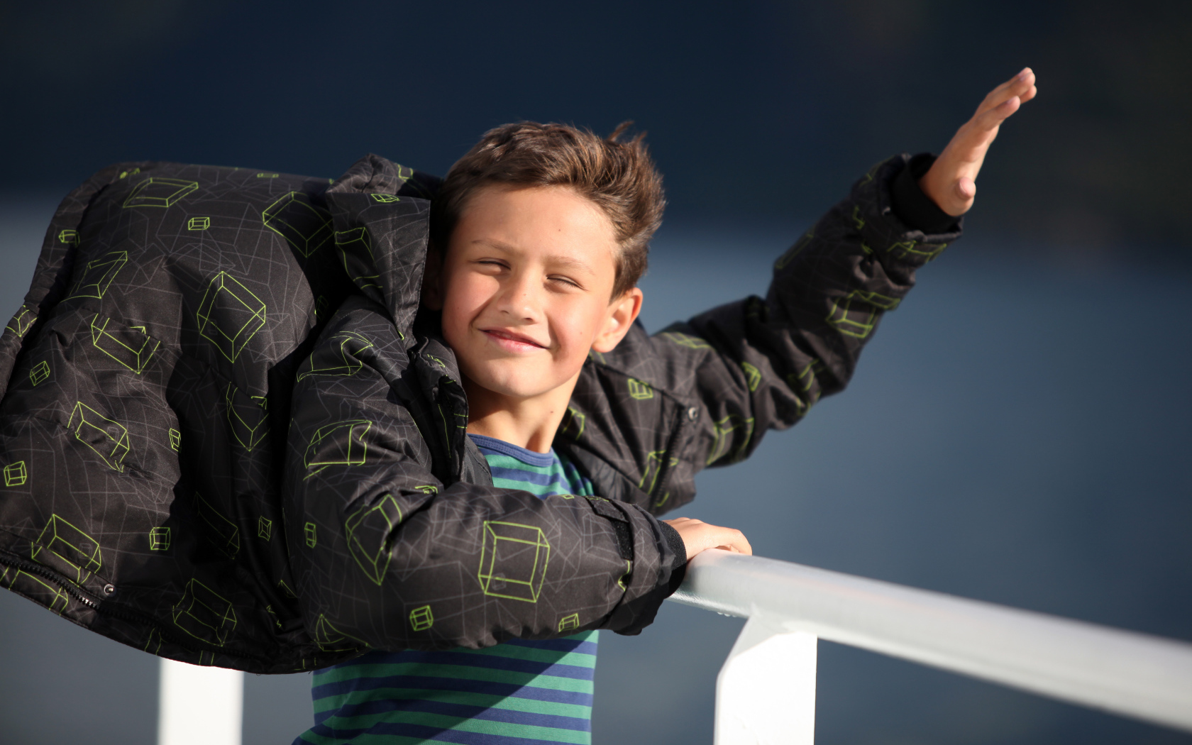 Веселый мальчик в куртке стоит на ветру в лучах солнца