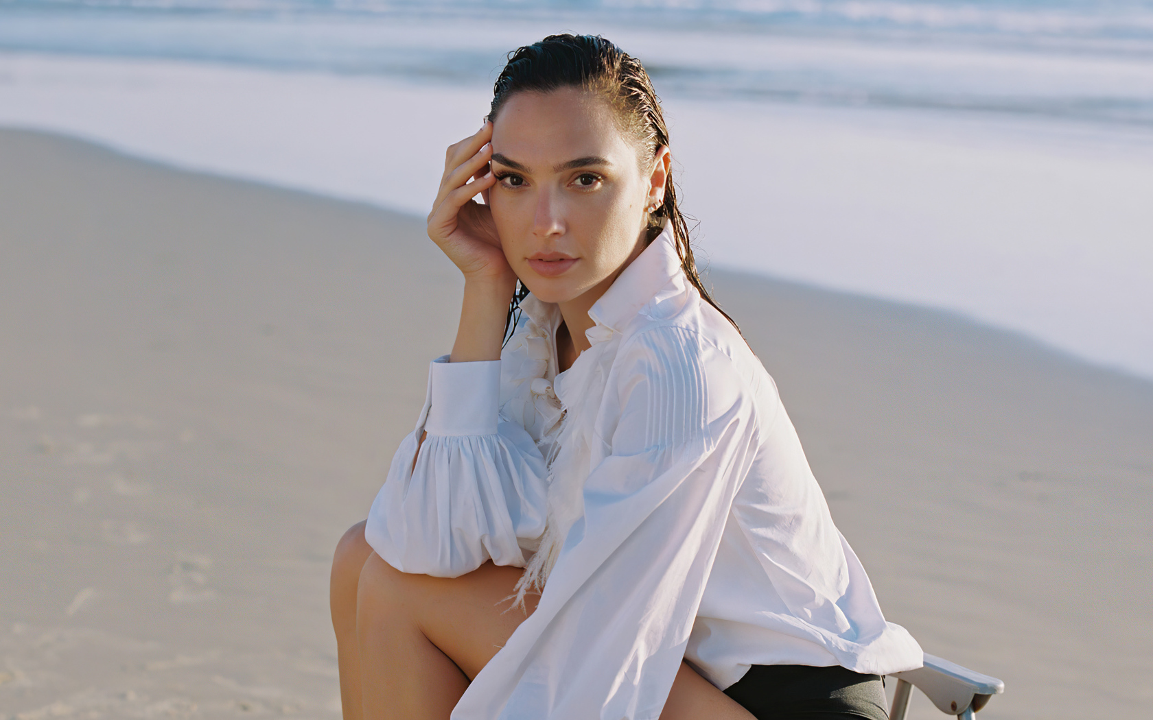 Актриса Галь Гадот в белой рубашке сидит на песке