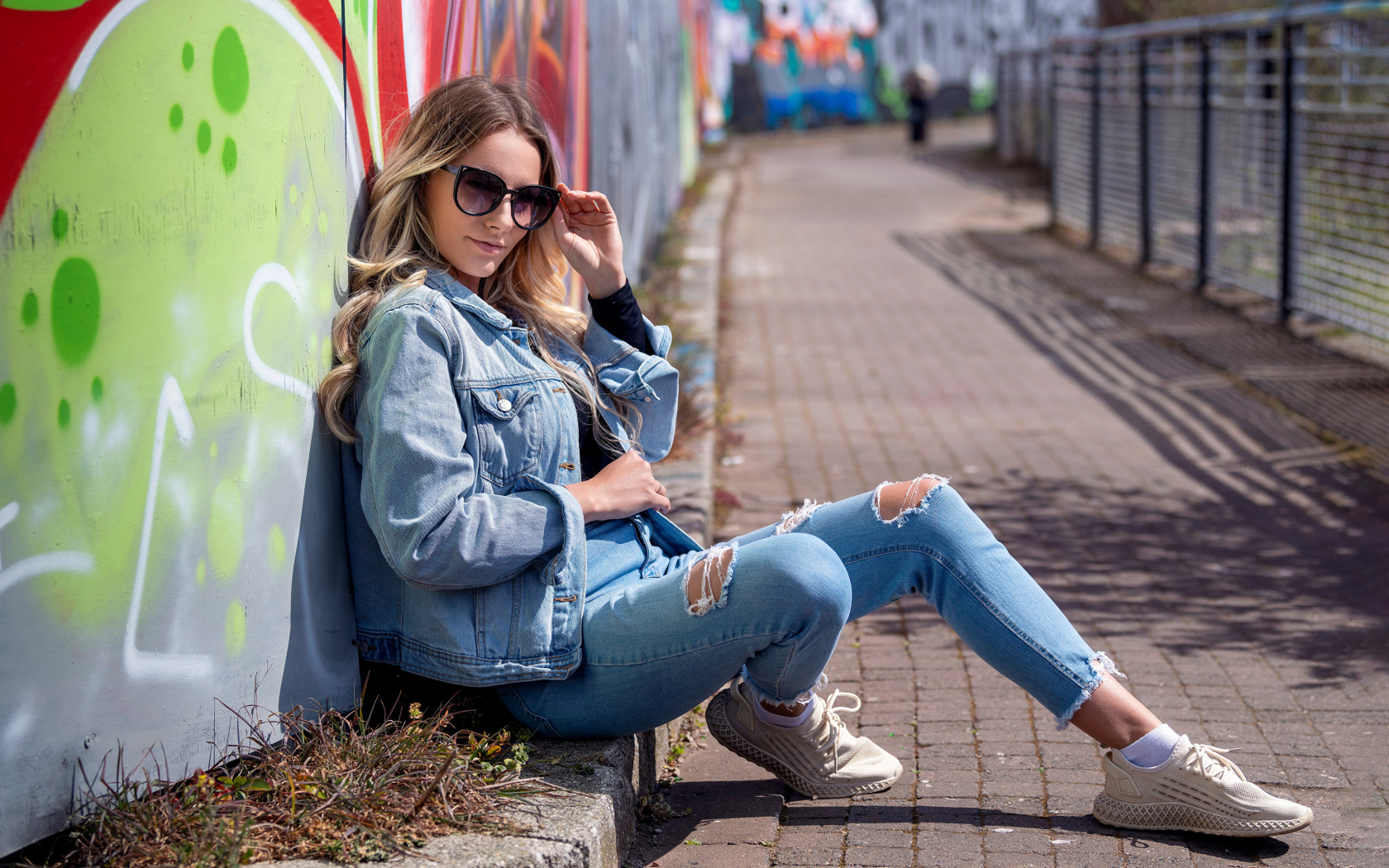 Красивая девушка в джинсовом костюме сидит у стены 