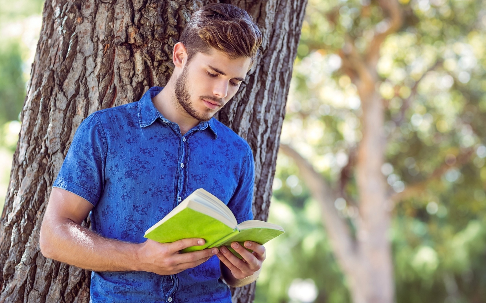 Красивый молодой мужчина стоит с книгой под деревом 