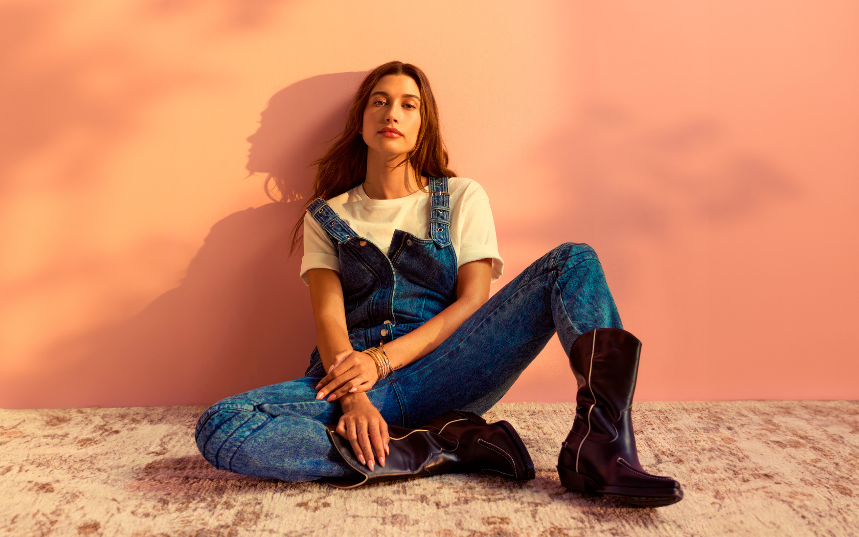 Американская модель Хейли Болдуин в джинсовом комбинезоне сидит у стены
