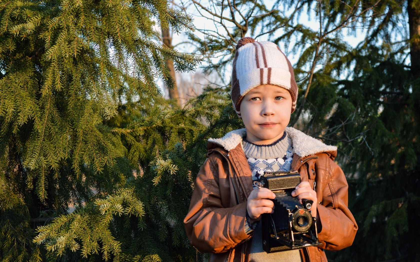 Мальчик с фотоаппаратом стоит у ели