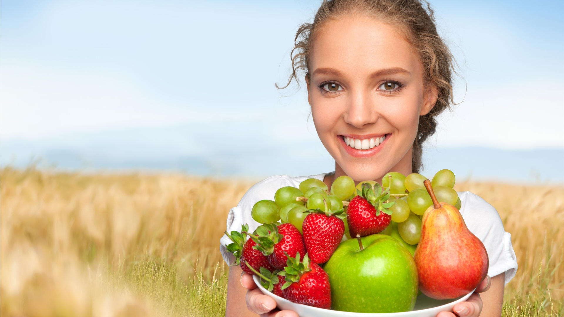 Улыбающаяся девушка шатенка с тарелкой с яблоками, виноградом, клубникой и грушей в руках
