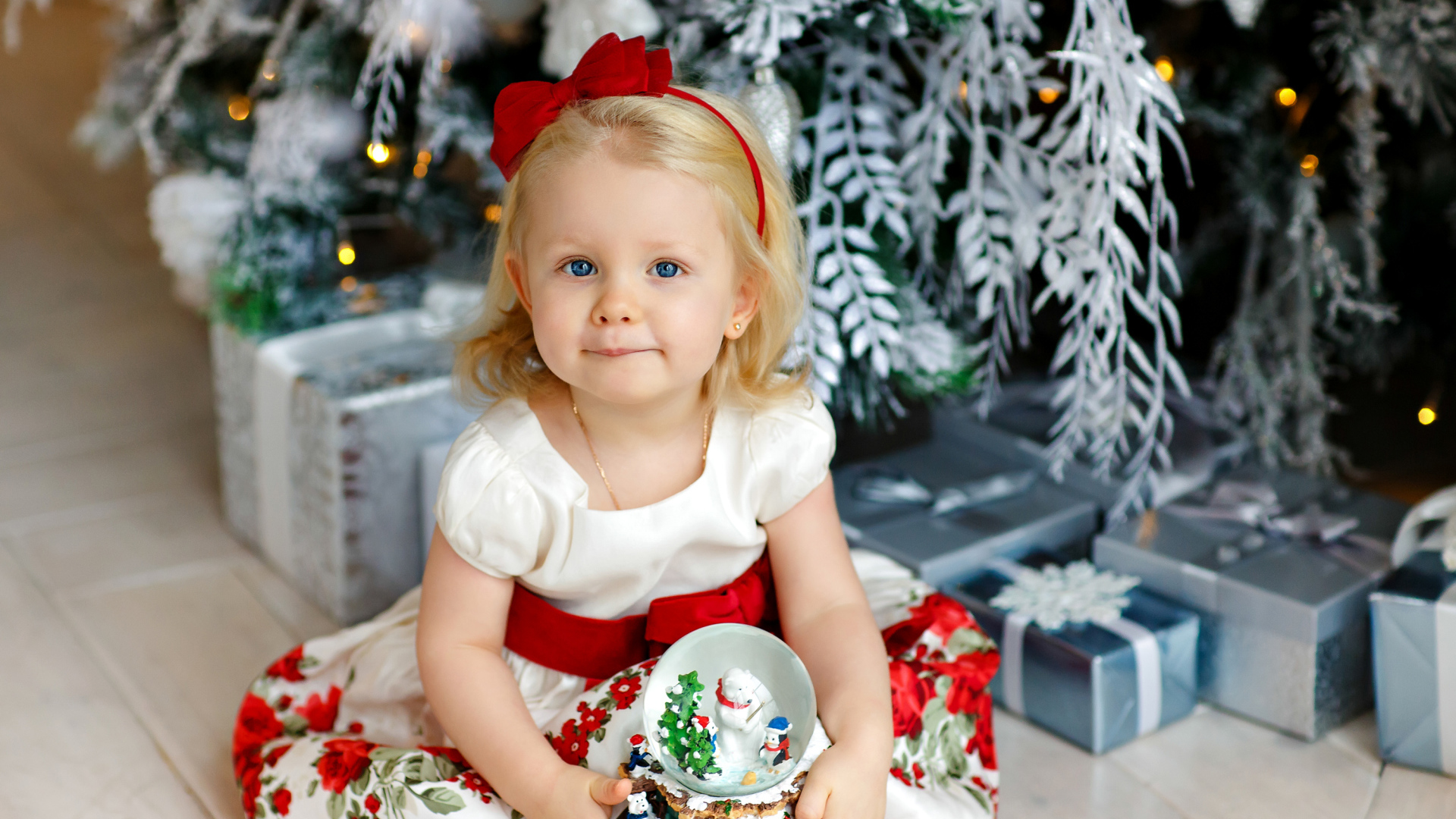 Маленькая девочка блондинка сидит с подарками под новогодней елкой