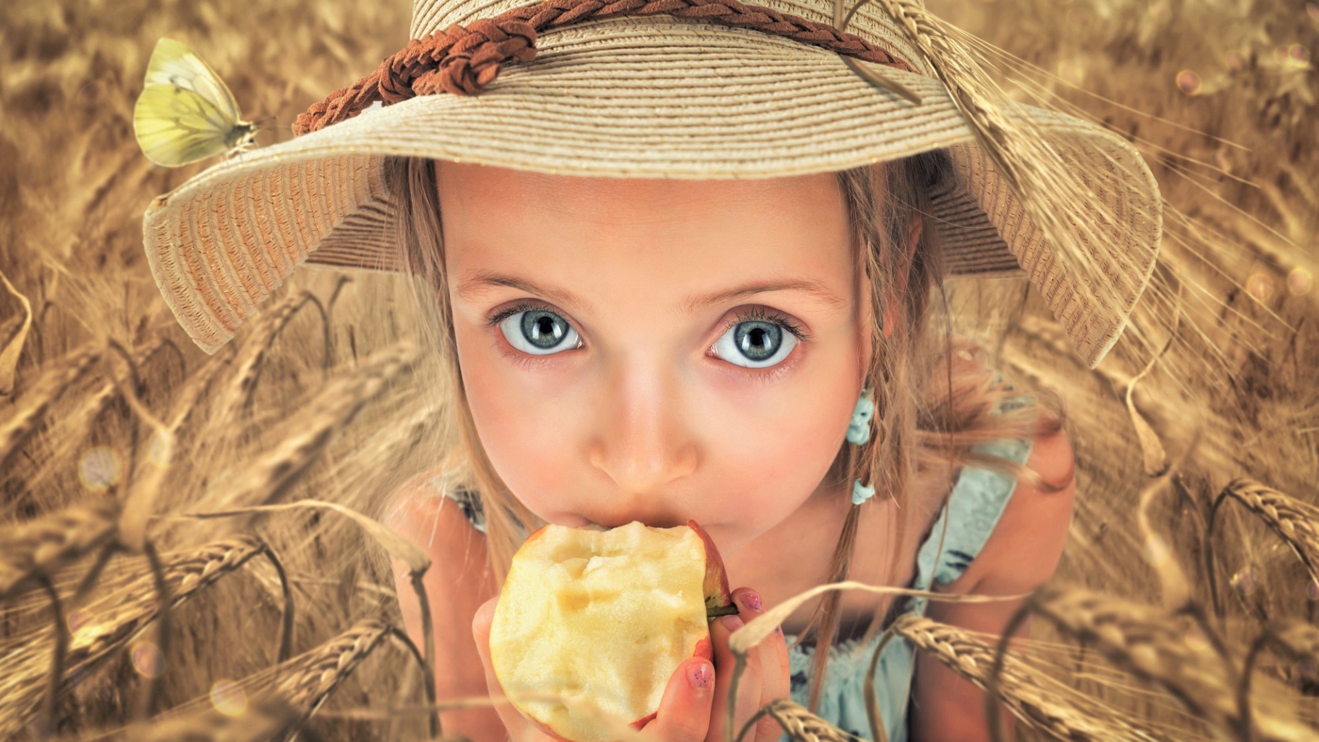 Маленькая голубоглазая девочка в шляпе ест яблоко